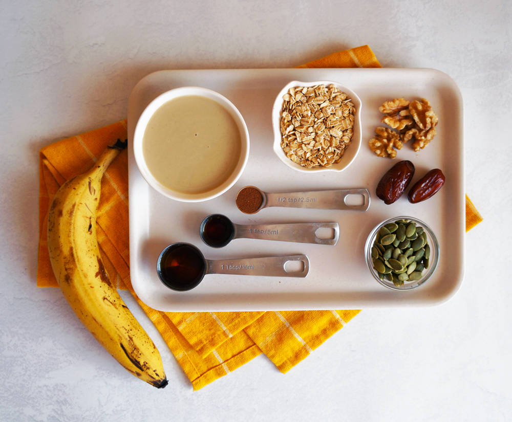 Banana bread granola ingredients; banana, oats, tahini, walnuts, pumpkin seeds, dates, cinnamon, maple syrup, and vanilla extract.