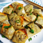Vegan mushroom cream cheese wontons on a white plate topped with black sesame seeds and chopped scallions with a side of sweet and sour sauce.