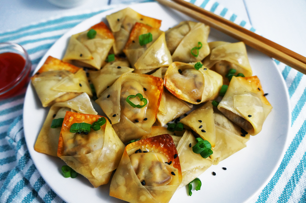 Vegan mushroom cream cheese wontons on a white plate topped with black sesame seeds and chopped scallions with a side of sweet and sour sauce.