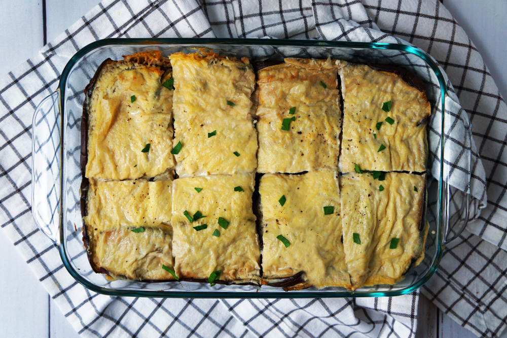 A glass tray of vegan moussaka with creamy bechamel sprinkled with parsley.