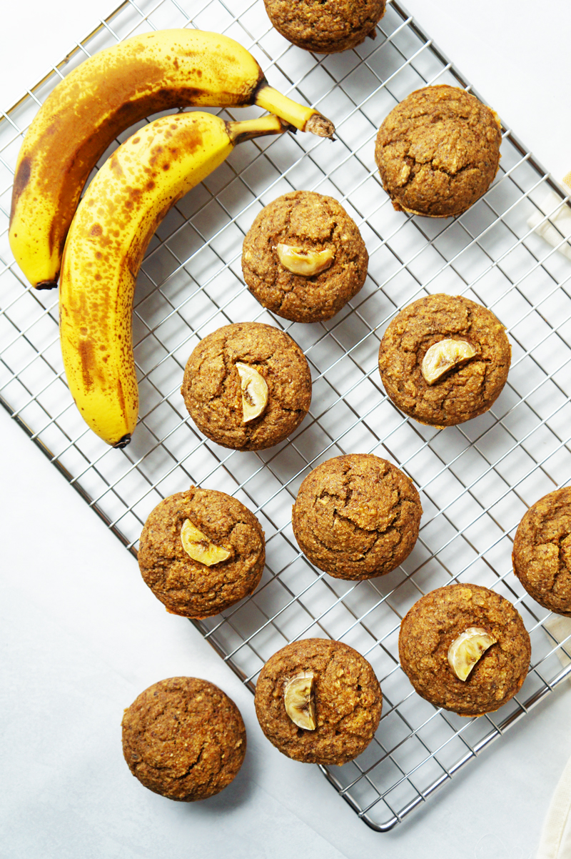 A dozen healthy, vegan, gluten-free banana bread muffins on a cooling rack with a bunch of ripe, spotty bananas nearby.