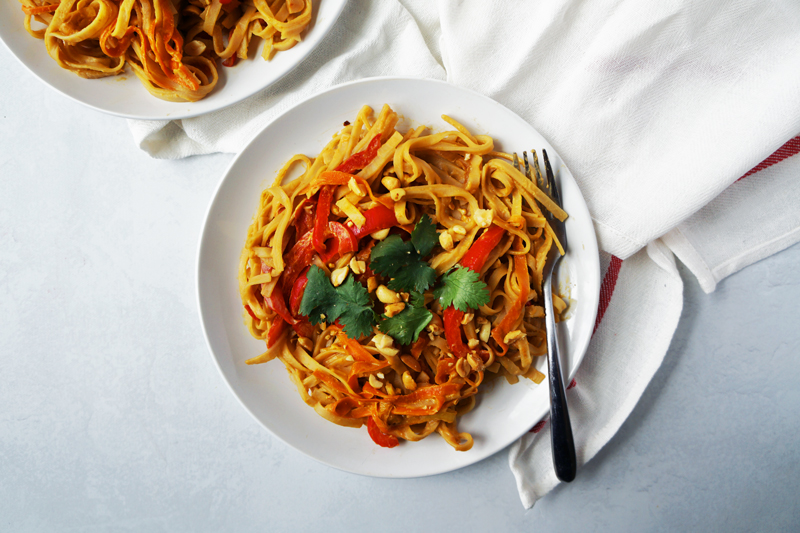 A plate of easy vegan peanut noodles with rice noodles, a soy peanut sauce, ribboned carrots, julienned red bell peppers, chopped peanuts, and cilantro.