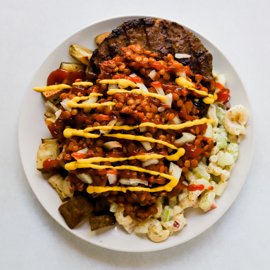 Plant-based garbage plate topped with ketchup, mustard, and onions on a white plate. 