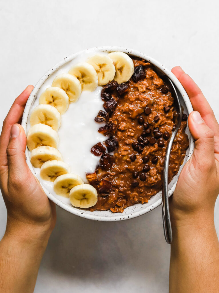 Bowl of vegan, gluten-free chocolate zucchini oatmeal topped with sliced bananas, dates, mini vegan chocolate tips, and coconut yogurt.