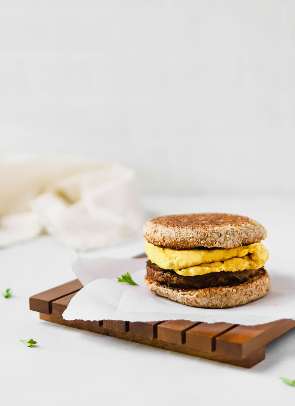 Breakfast sandwich with folded vegan egg, vegan sausage patty, and toasted english muffin on a piece of white parchment paper. 
