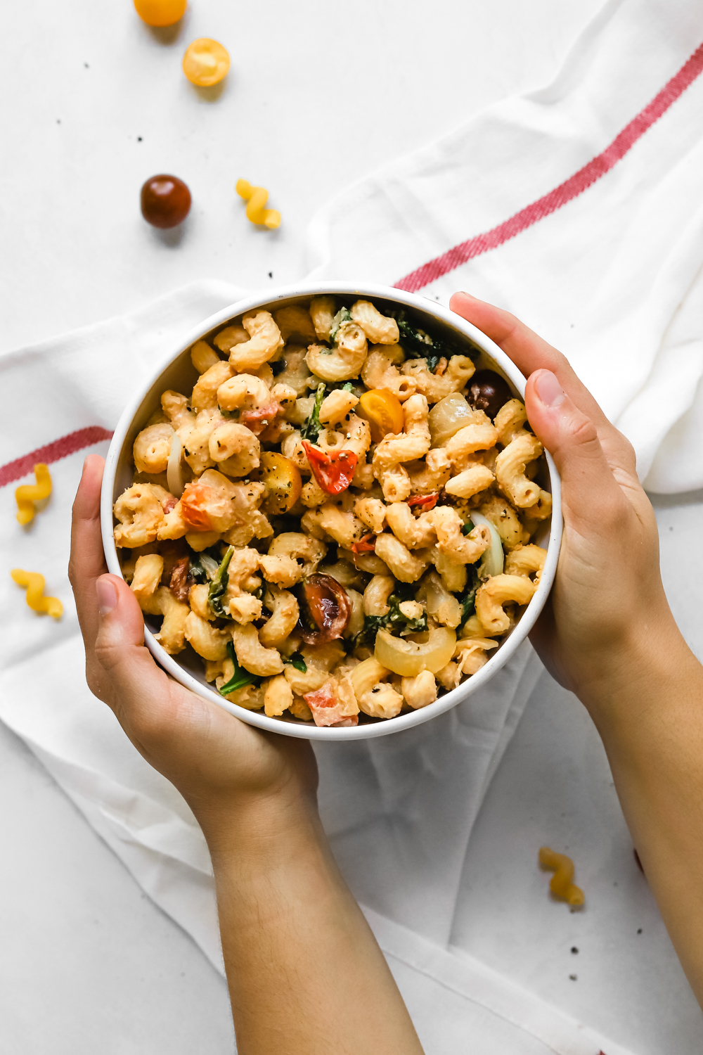 Hands holding a bowl of vegan hummus pasta with veggies.