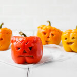 Spooky vegan bell pepper jack o'lanterns smiling at the camera on a white wooden table.