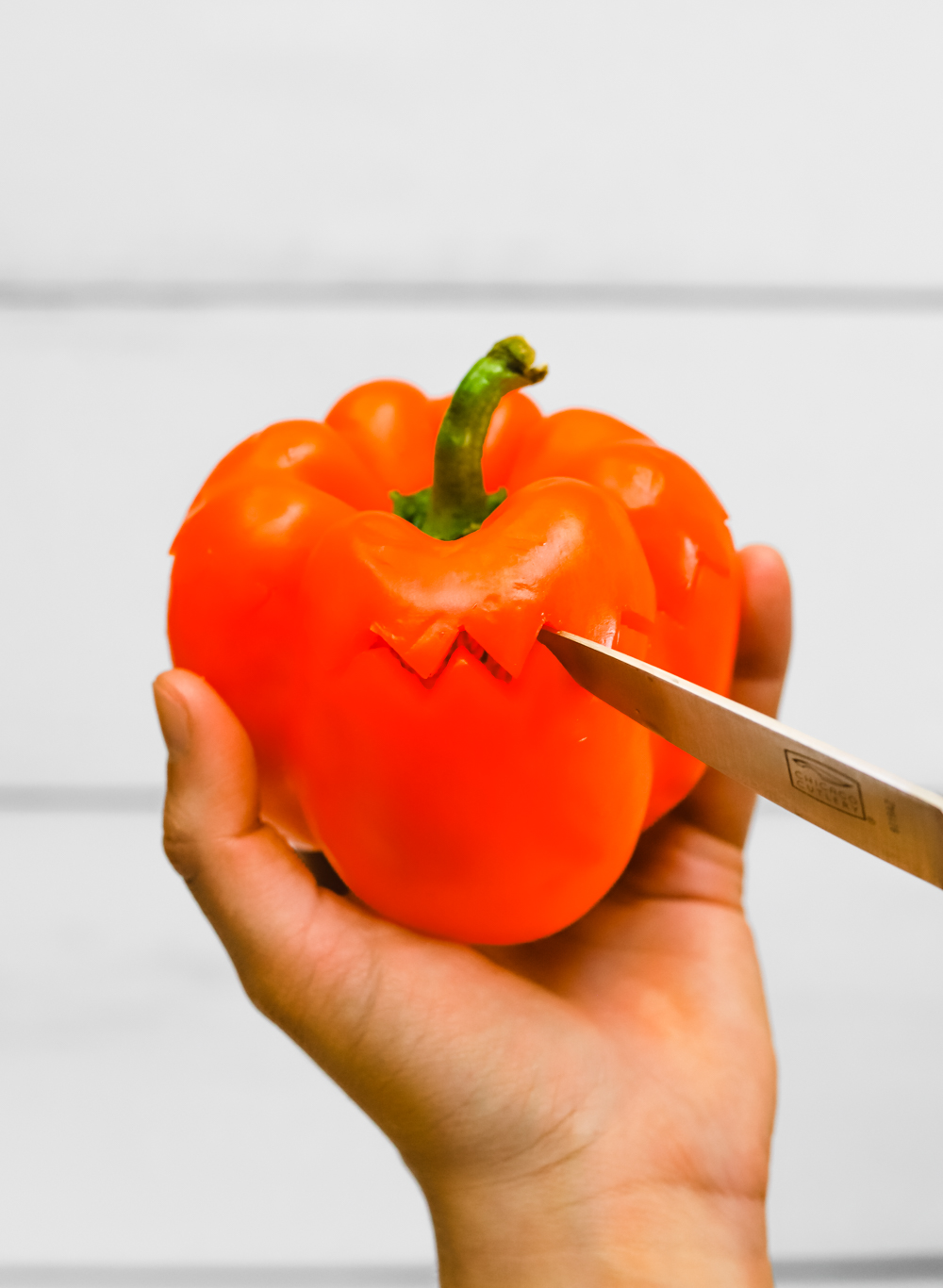Pairing knife cutting the top off of an orange bell pepper in a zig zag design to make a bell pepper jack o'lantern.