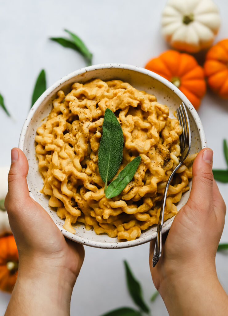 Hands holding a bowl of vegan, gluten-free pumpkin miso pasta with cracked black pepper and a fresh sage leave for garnish.