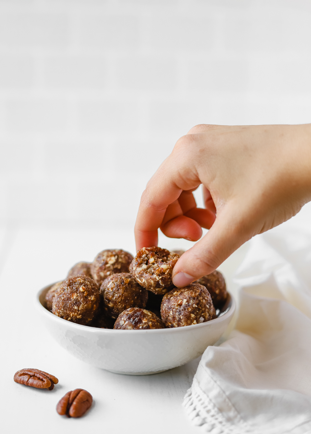 Hand grabbing a pecan pie bliss ball from a small white bowl.