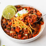 Bowl of hearty vegetarian tvp chili, topped with vegan chili and sour cream, chopped green onions, and lime wedges.