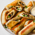 A plate of vegan philly cheesesteak egg rolls with a cheese dipping sauce on a white plate