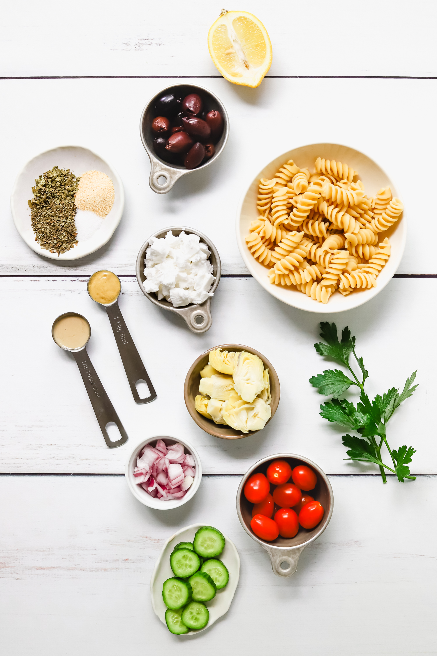 Ingredients for greek pasta salad: pasta, kalamata olives, cucumber, tomatoes, artichoke hearts, red onions, parsley, and vegan feta