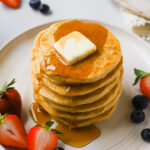 a tall stack of vegan protein pancakes with butter and maple syrup surrounded by berries